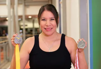 A woman with dark hair pulled back in workout clothes is holding two medals up and smiling at the camera. There is a gymnasium in the background. 