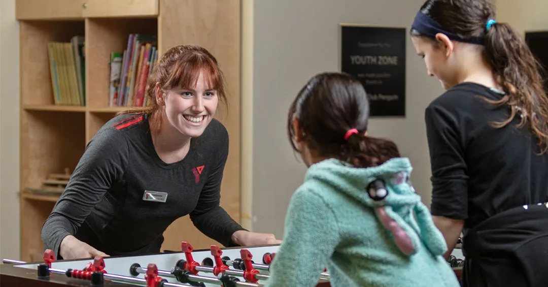 A young woman in a Y uniform shirt is playing foosball with two younger girls with their backs to the camera. She is smiling at the young girls and positioned for action with her hands on the handles. 