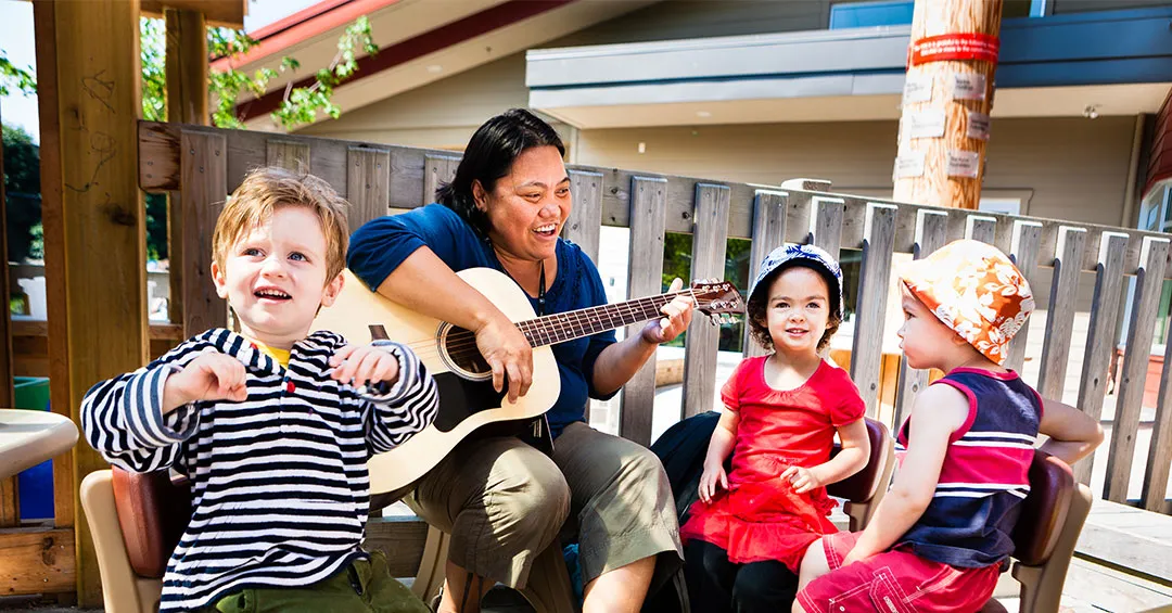 A middle aged woman is sitting in a chair on a balcony outside on a sunny day. She's holding a guitar and interacting with three young children sitting around her.